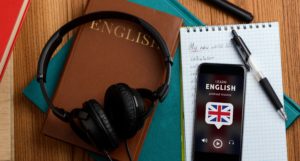 Un casque avec un portable posés sur une table et des cahiers. Sur le portable on retrouve la phrase "Learn English" pour rappeler la formation anglais à distance