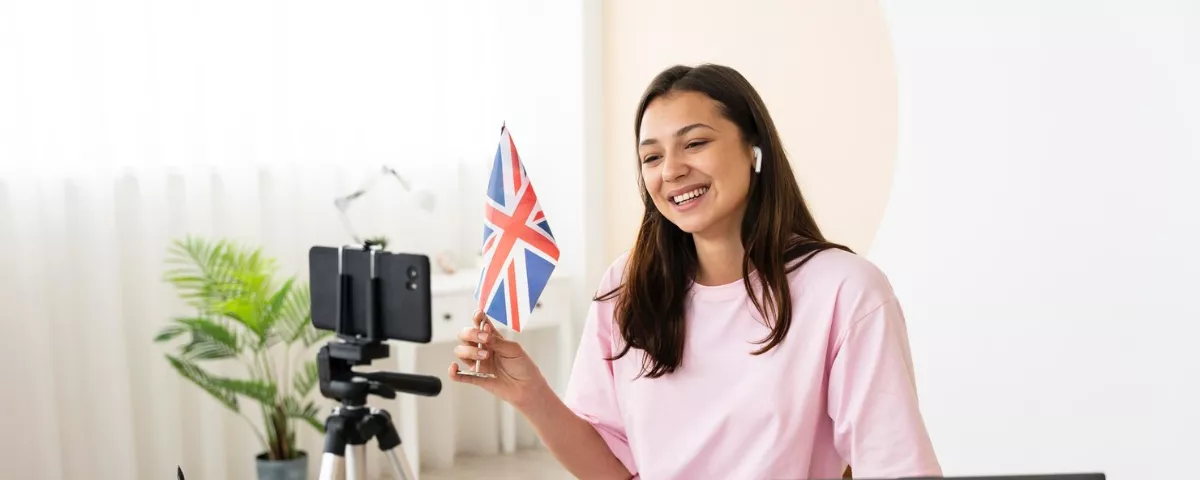 Une femme souriante tenant un drapeau du Royaume-Uni face à un smartphone sur trépied pour un cours de langue en ligne.