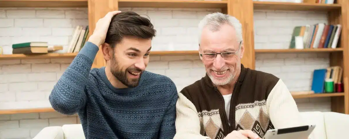 Deux professionnels, un homme plus jeune et un homme plus mûr, assis côte à côte et souriants, analysant ensemble des données de reporting ou une formation sur une tablette numérique.