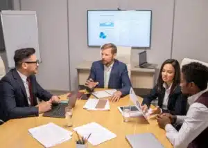 Équipe de professionnels en réunion de travail autour d'une table avec graphiques de performance sur écran.