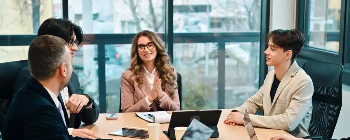 Quatre collègues de bureau en réunion collaborative autour d'une table avec des tablettes et des documents, applaudissant une présentation réussie dans une salle de conférence lumineuse.