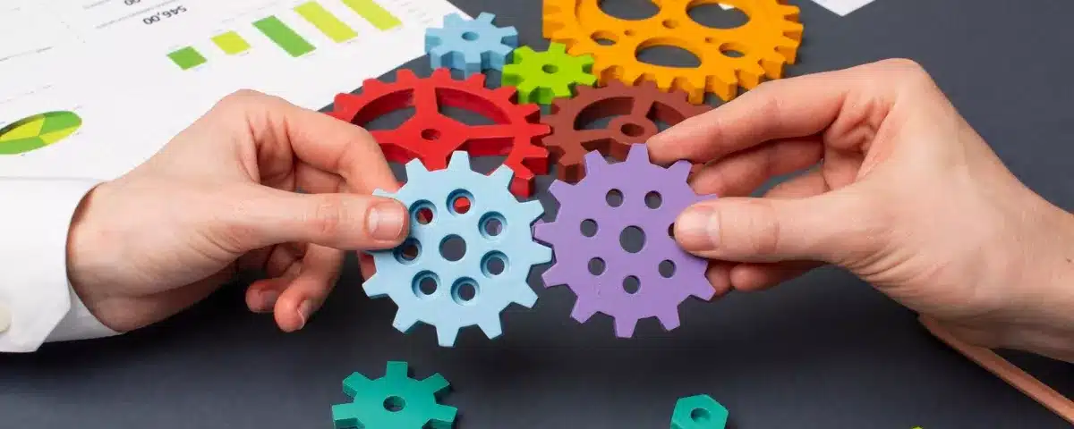 Mains d'une équipe de travail unissant des engrenages de couleur sur un bureau couvert de graphiques d'analyse commerciale.