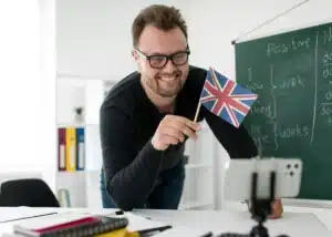 Formateur d'anglais souriant tenant un petit drapeau britannique lors d'une visioconférence professionnelle en entreprise.